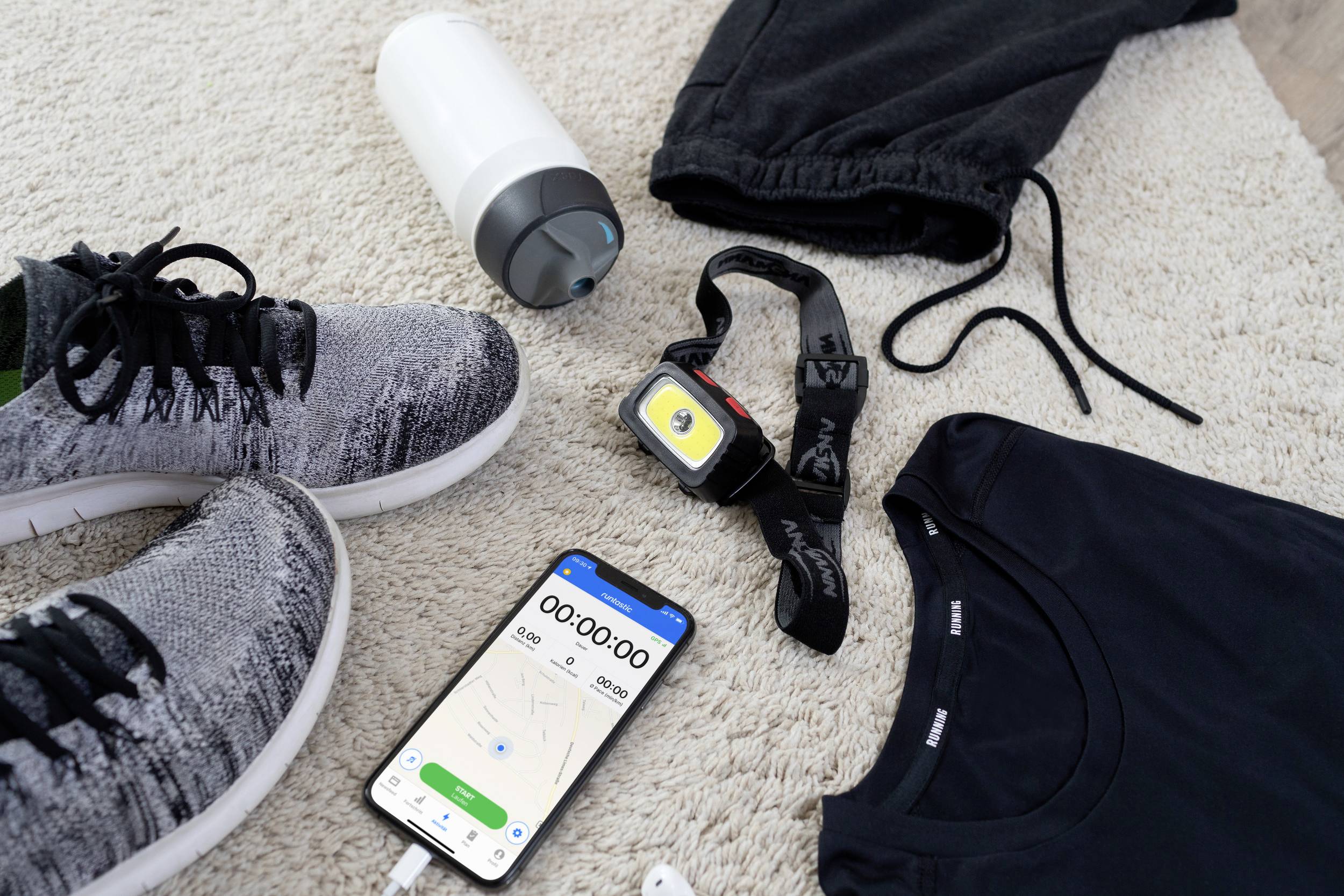 Sportausrüstung liegt auf einem Teppich: Laufschuhe, Sportkleidung, Wasserflasche, Fitness-Armbanduhr und ein Smartphone mit einer Stoppuhr-App.