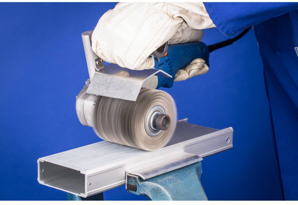 Eine Person schleift mit einer Schleifmaschine ein Metallstück. Sie trägt Arbeitshandschuhe; der Hintergrund ist blau.