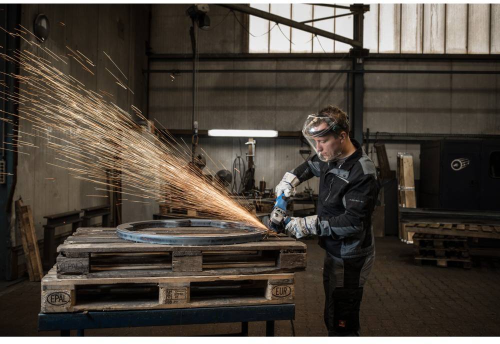 Ein Arbeiter in Schutzkleidung schleift ein großes Metallteil auf einer Palette in einer Werkstatt, Funken fliegen umher.