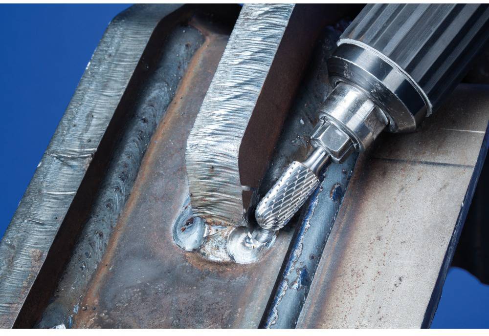 Nahaufnahme eines Metallarbeiters, der mit einem Fräswerkzeug Schweißnähte glättet. Sichtbare Metallschicht und blauer Hintergrund.