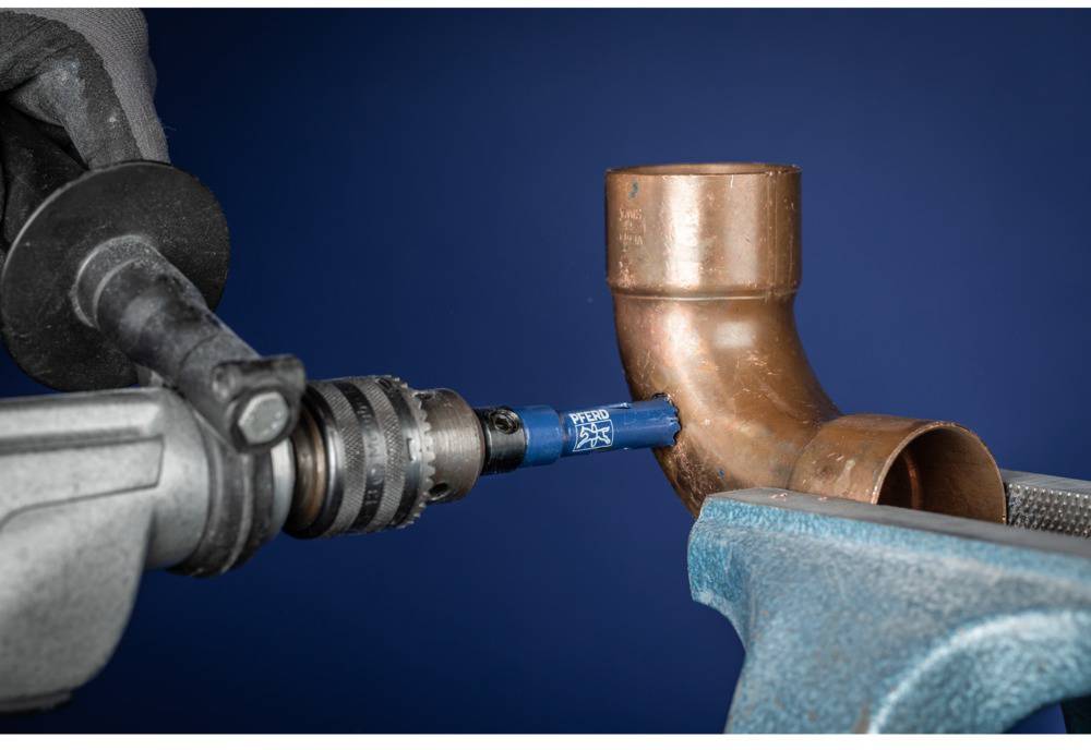 Hand hält Bohrmaschine mit Aufsatz, der Kupferrohr in Schraubstock schneidet; symbolisiert Handwerk oder Installationstechnik.
