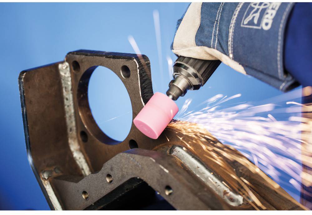 Eine Hand in einem Arbeitshandschuh schleift ein Metallteil mit einer elektrischen Schleifmaschine, Funken sprühen.