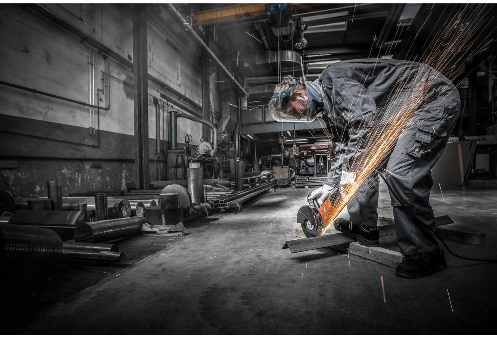 Ein Arbeiter in Schutzkleidung schleift Metall in einer Fabrik, Funken sprühen. Die Umgebung zeigt Werkzeuge und industrielle Maschinen.