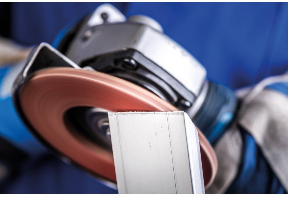 Ein Arbeiter schleift ein Metallstück mit einer elektrischen Schleifmaschine, Funken fliegen. Sicherheitshandschuhe sichtbar.