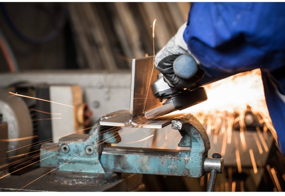 Eine Person schleift Metall mit einem Winkelschleifer in einer Werkstatt. Funken fliegen vom Metallstück weg.