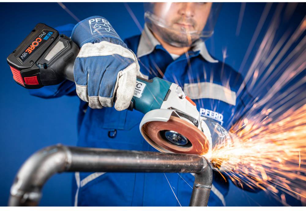 Ein Arbeiter mit blauer Uniform schleift mit einer elektrischen Schleifmaschine ein Rohr. Funken fliegen von der Schleifscheibe.