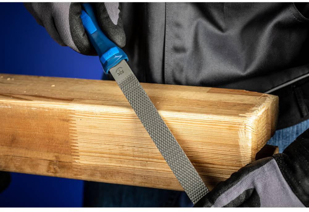 Eine Person feilt ein Stück Holz mit einer blauen Feile. Sie trägt schwarze Handschuhe und einer schwarzen Jacke.