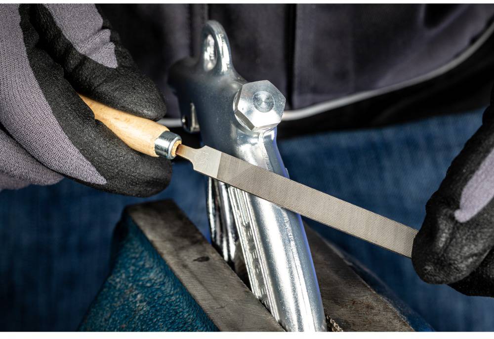 Nahaufnahme einer Person, die mit Handschuhen und einer Feile ein Metallstück bearbeitet, das in einem Schraubstock eingespannt ist.