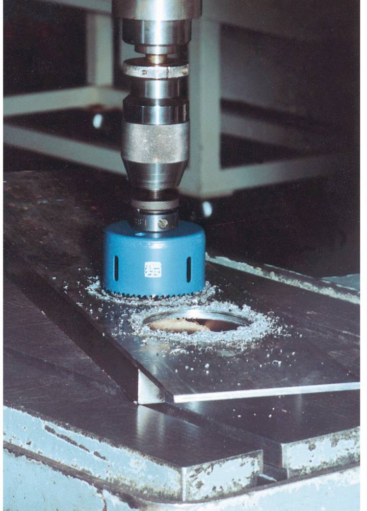 Eine blaue Lochsäge ist an einer Bohrmaschine befestigt und bohrt ein rundes Loch in eine Metallplatte auf einem Arbeitstisch.