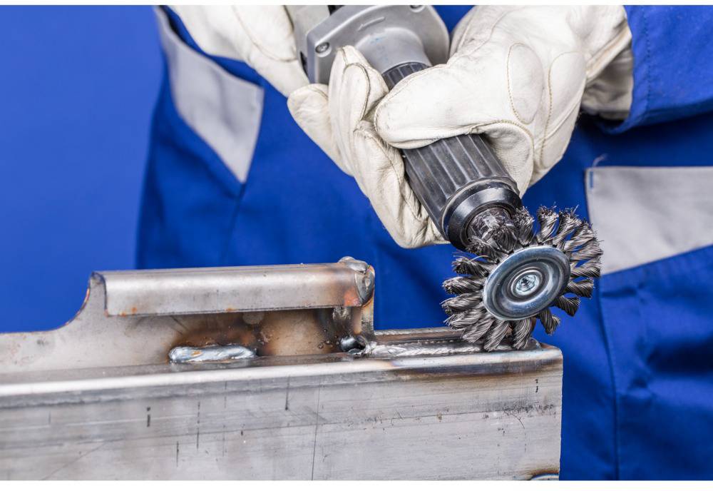 Eine Person in Schutzkleidung reinigt mit einer Drahtbürste ein Metallstück. Das Metall zeigt Spuren von Schweißarbeiten.