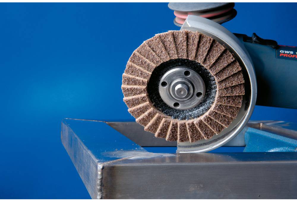 Eine Schleifmaschine mit einer Schleifscheibe aus Schaumstoff bearbeitet die Kante eines Metallstücks vor blauem Hintergrund.