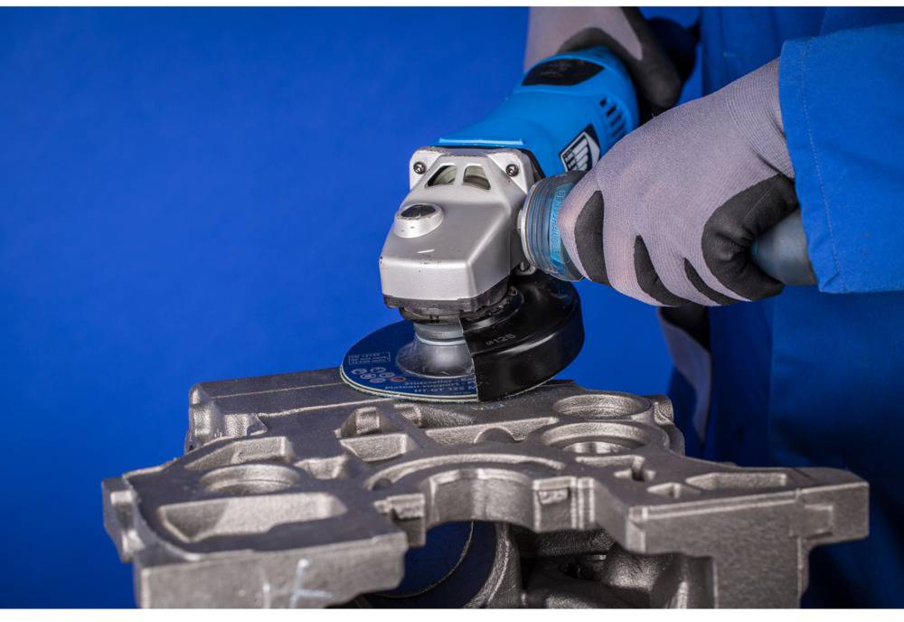 Person in Arbeitskleidung schleift Metallstück mit einer blauen Winkelschleifmaschine vor blauem Hintergrund.