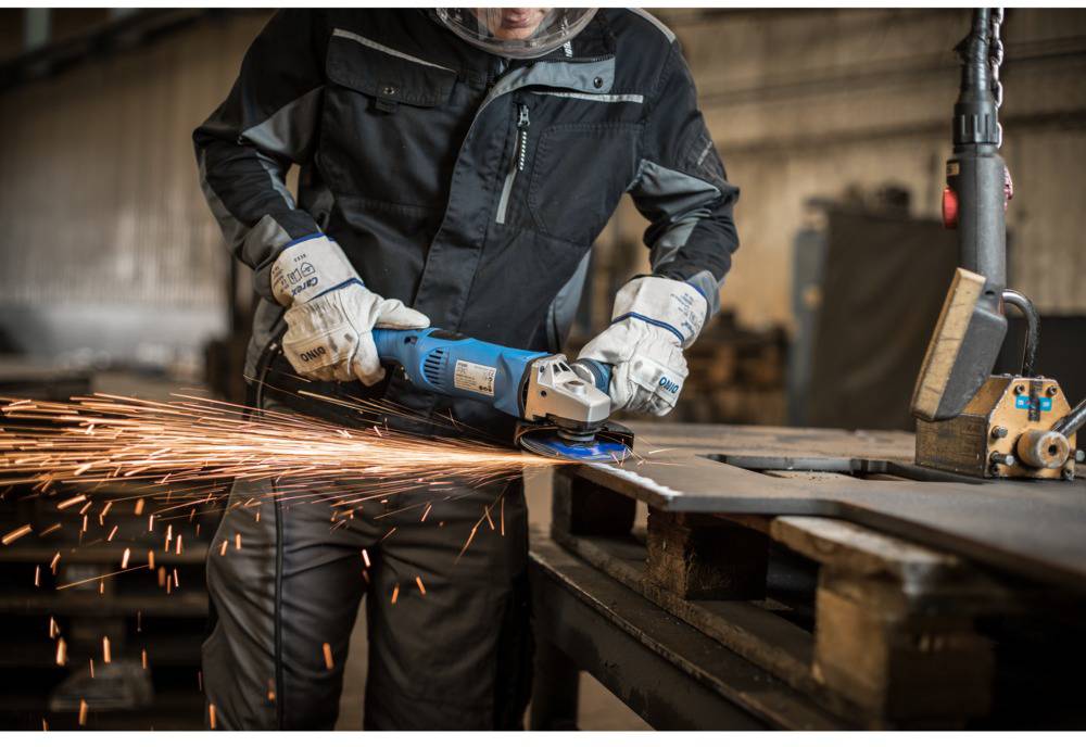 Ein Arbeiter schleift Metall mit einer Schleifmaschine, Funken fliegen. Er trägt Schutzkleidung und arbeitet in einer Werkstatt.
