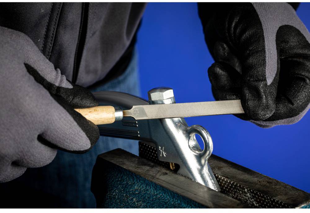 Eine Person feilt einen Schraubenschlüssel in einem Schraubstock mit einem Metallfeilenwerkzeug, trägt dabei schwarze Schutzhandschuhe.