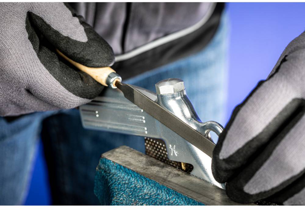 Eine Person mit schwarzen Handschuhen schärft eine Klinge mit einem kleinen Schleifwerkzeug. Die Klinge liegt auf einer Metallhalterung.