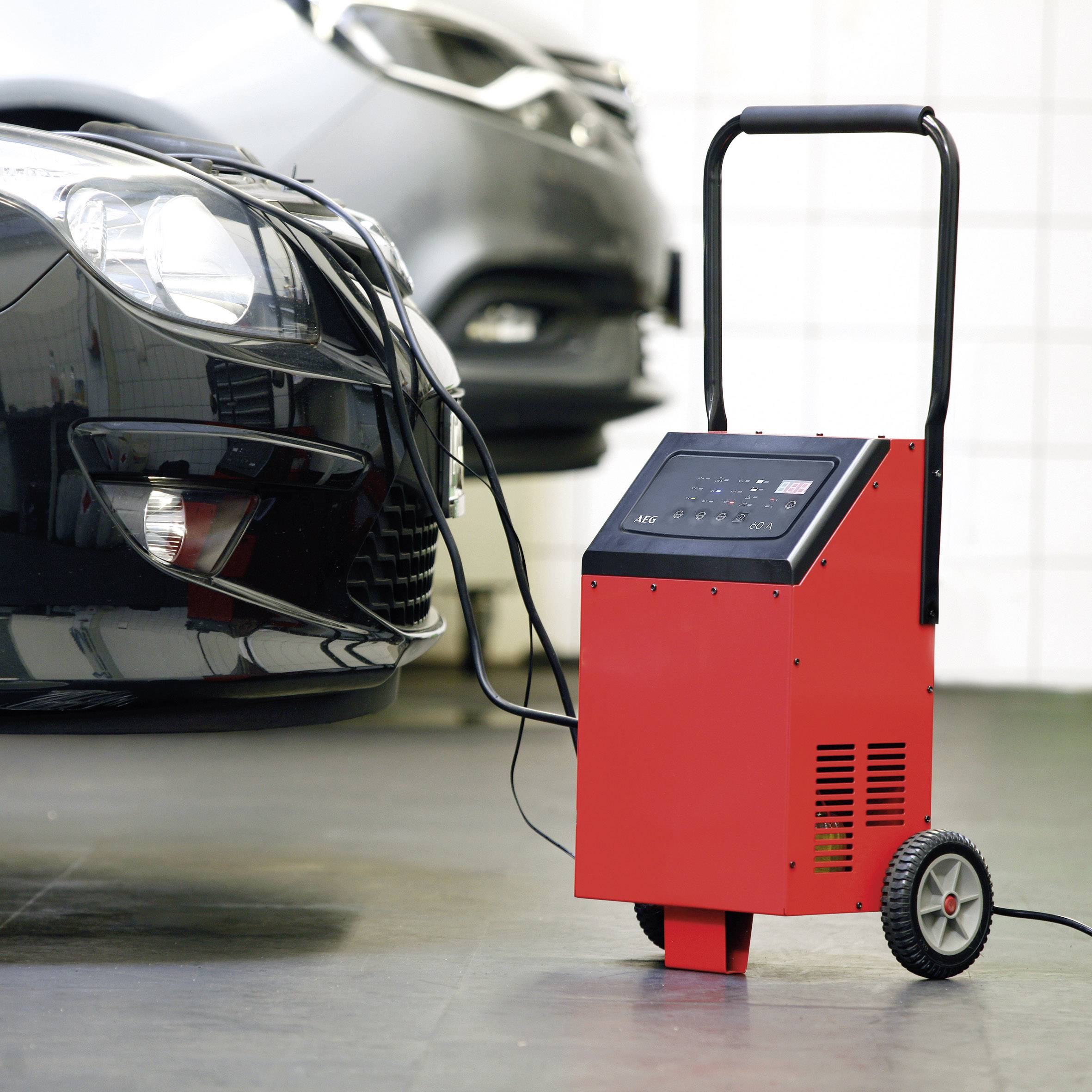 Ein schwarzes Auto wird in einer Garage mit einem roten Batterieladegerät aufgeladen. Das Ladegerät steht auf zwei Rädern.