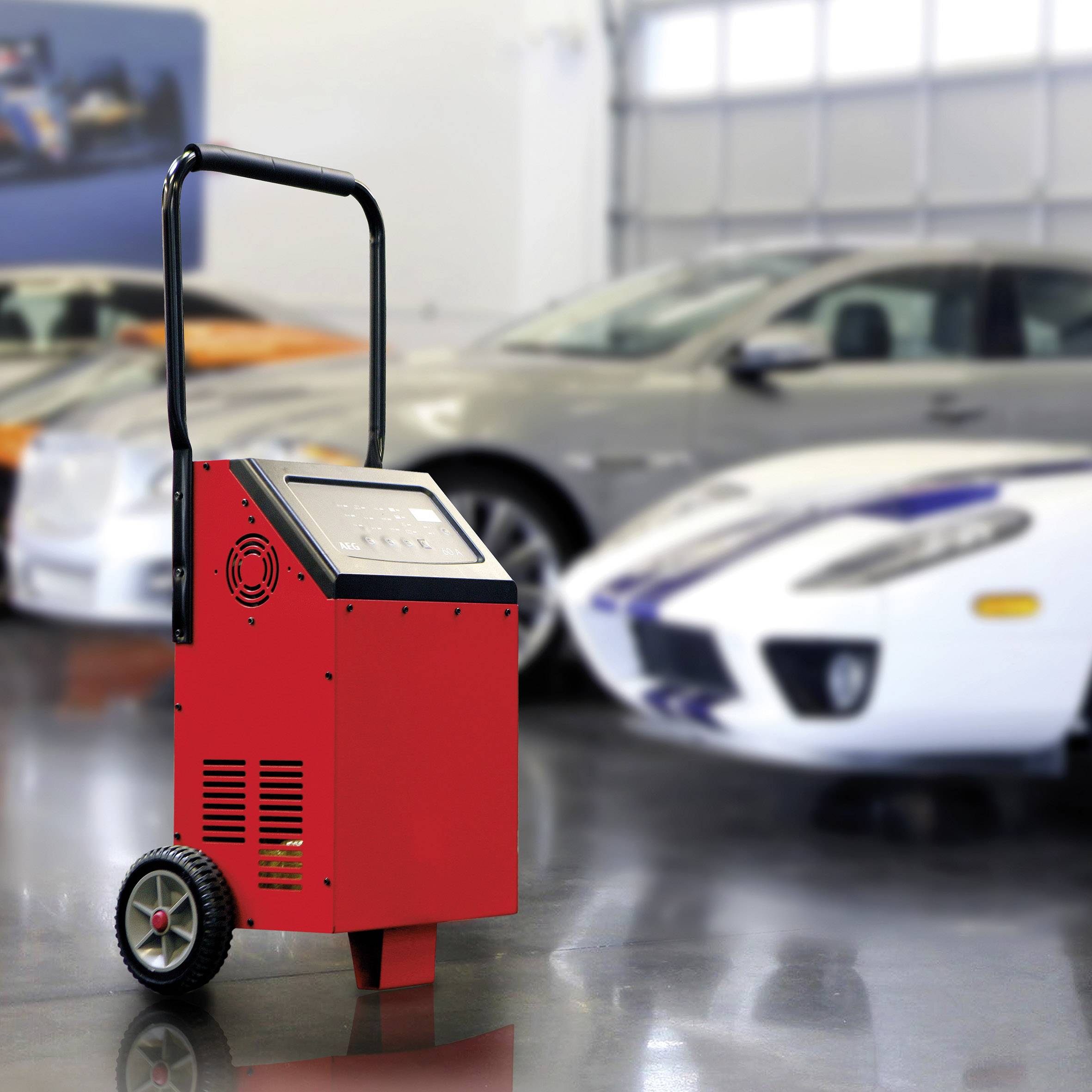 Ein rotes mobiles Batterieladegerät steht in einer Garage vor drei Autos. Im Hintergrund sind unscharf zwei Sportwagen erkennbar.