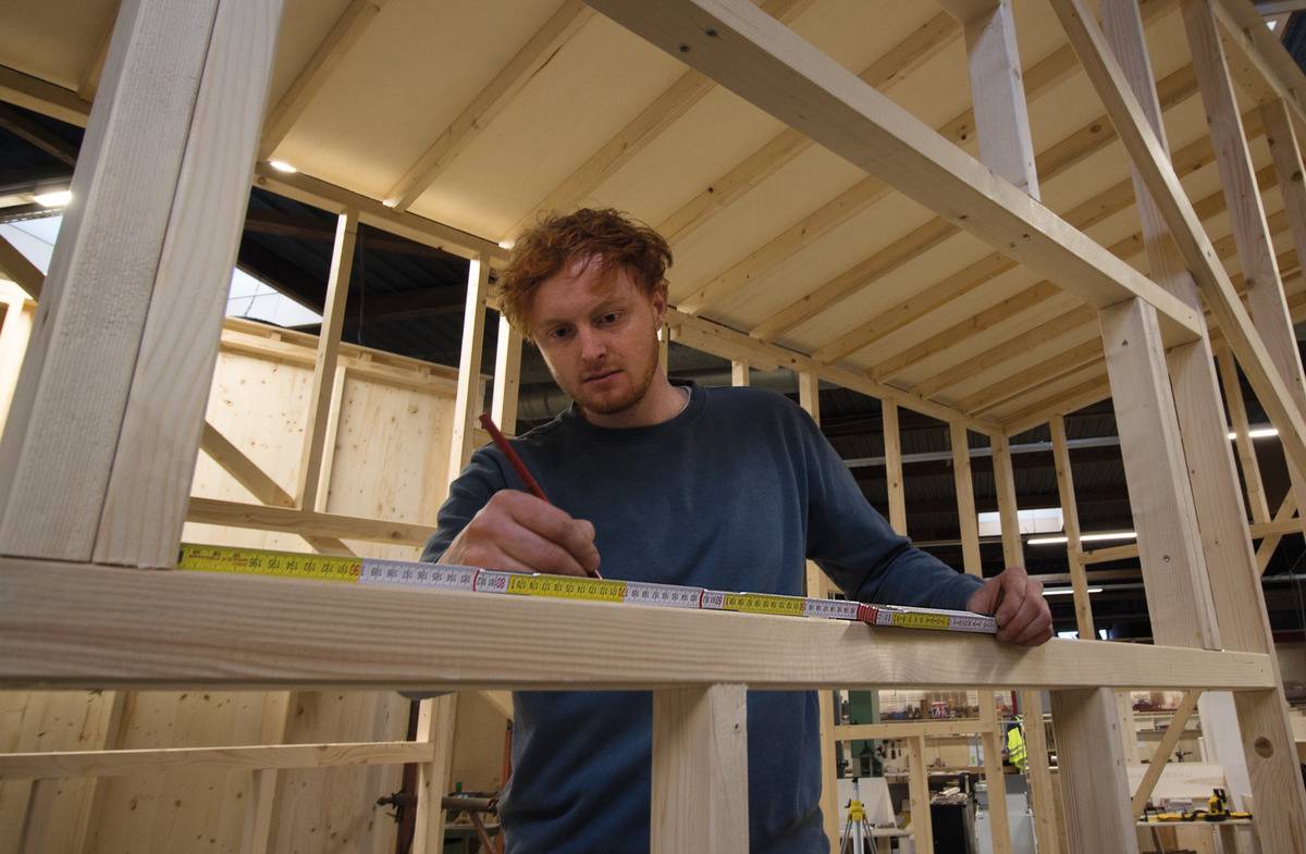 Ein Mann misst mit einem Maßband und notiert auf einem Holzrahmen in einer Werkstatt, konzentriert bei der Arbeit an einem Holzbauprojekt.