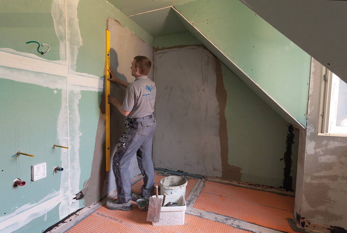 Ein Handwerker nivelliert eine Gipsplatte in einem Dachbodenausbau. Der Raum hat eine Schräge, und diverse Bauwerkzeuge sind sichtbar.
