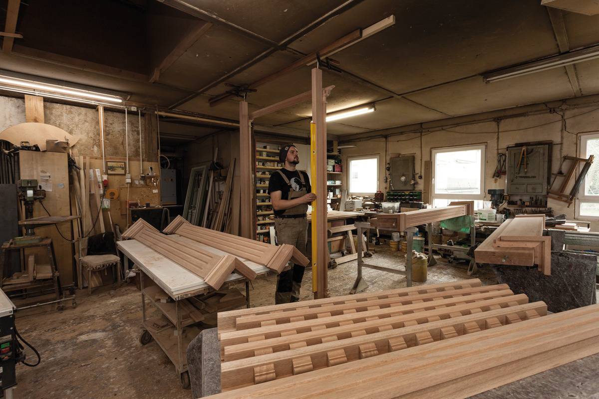 Eine Werkstatt mit einem Handwerker, der Holzarbeiten durchführt. Es sind verschiedene Holzstücke, Werkzeuge und Maschinen zu sehen.