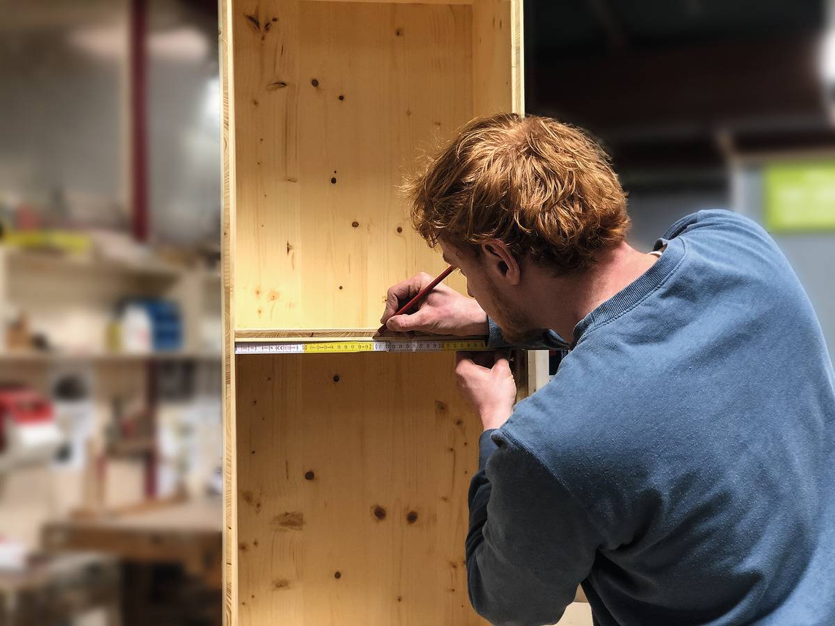 Eine Person misst ein Holzregal in einer Werkstatt mit einem Maßband und markiert die Stelle mit einem Bleistift.