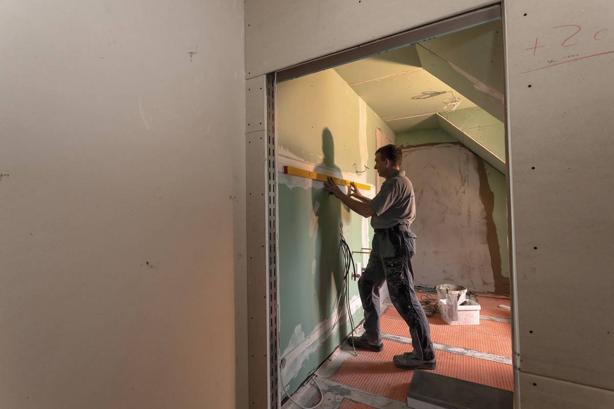 Ein Bauarbeiter prüft mit einer Wasserwaage die Ausrichtung einer Wand in einem unfertigen Raum. Der Boden ist mit Schutzmatten ausgelegt.
