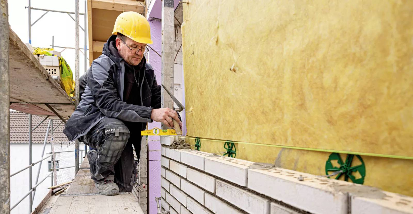 Ein Bauarbeiter mit gelbem Schutzhelm verwendet eine Wasserwaage, um Ziegelsteine an einer Wand mit Dämmung auszurichten, während er auf einem Gerüst steht.