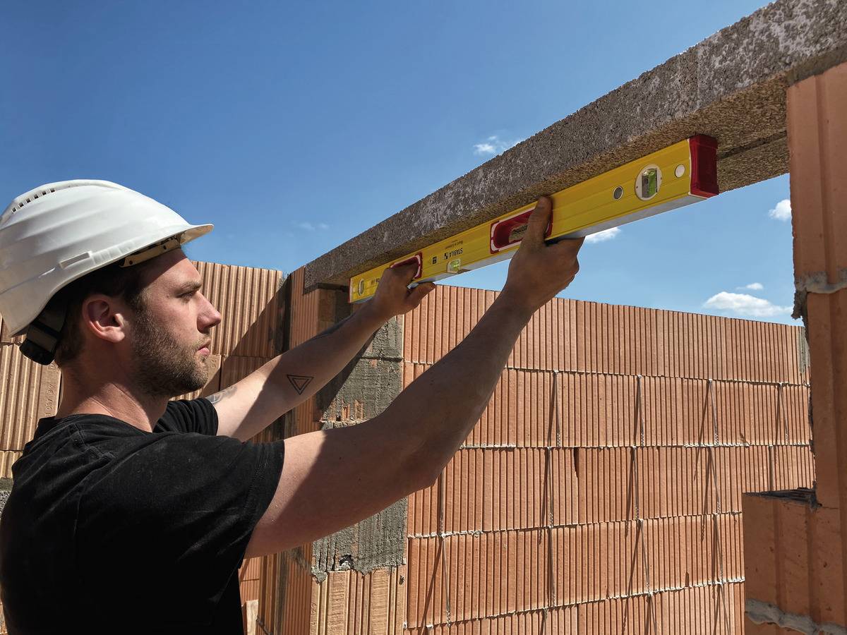 Ein Arbeiter mit Helm überprüft mit einer Wasserwaage die horizontale Ausrichtung einer Mauer aus Ziegelsteinen unter blauem Himmel.