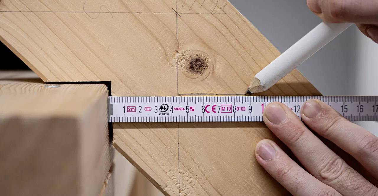 Eine Hand hält einen Maßstab und einen Stift, um Holz für einen präzisen Schnitt zu markieren, auf einer Orientierungslinie.
