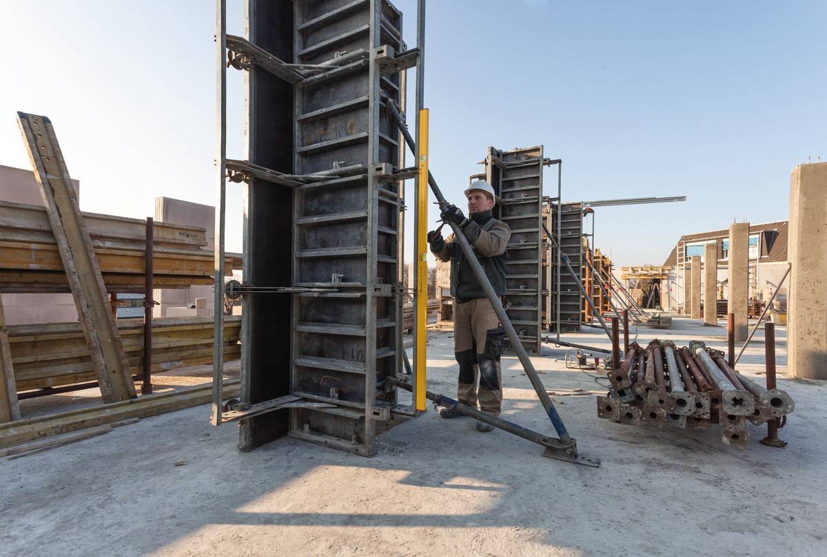 Ein Bauarbeiter baut auf einer Baustelle Gerüste auf. Um ihn herum sind Stahlträger und andere Baugeräte. Es ist sonnig.