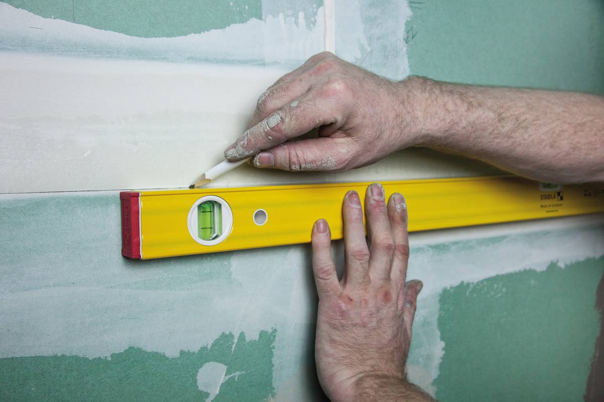 Eine Person markiert mit einem gelben Wasserwaage eine gerade Linie auf einer grünen Trockenbauwand.
