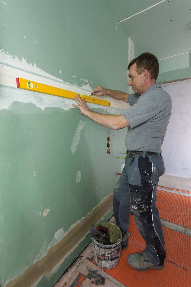 Mann in Arbeitskleidung richtet eine Wasserwaage an einer grün gestrichenen Wand aus. Im Hintergrund sind Werkzeuge und Baumaterialien sichtbar.