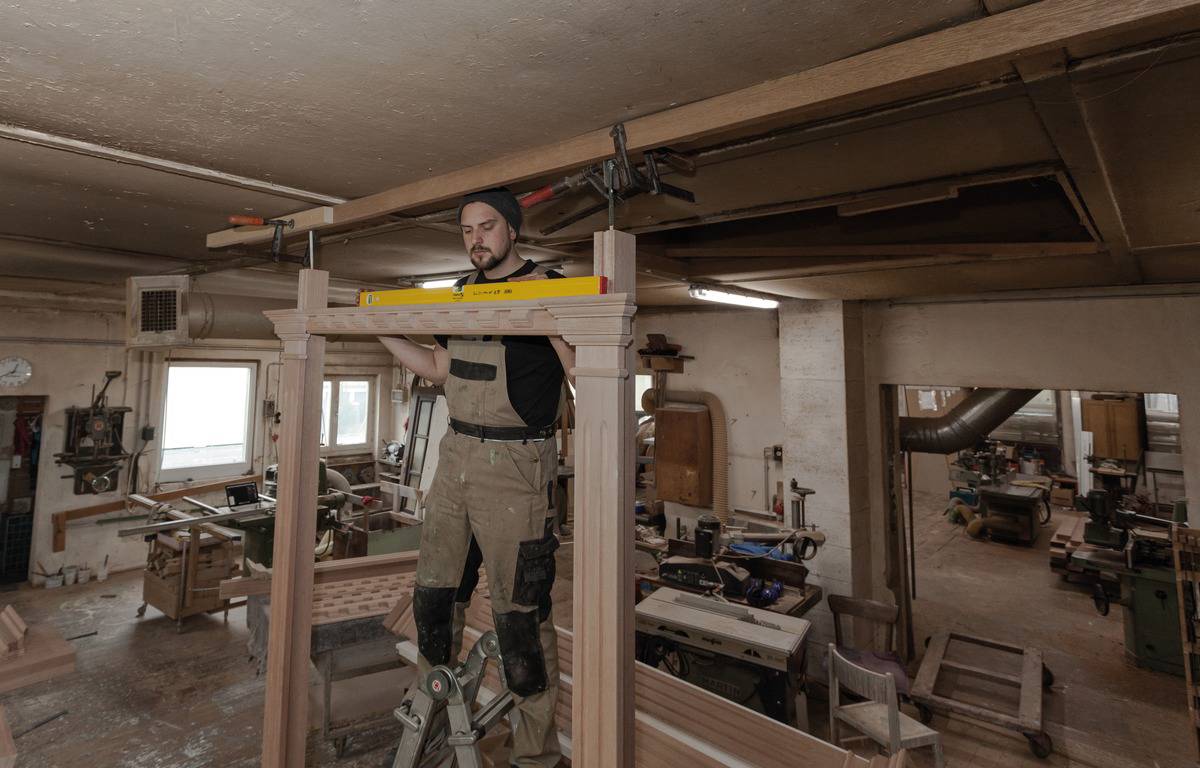 Ein Handwerker steht auf einer Leiter und verwendet eine Wasserwaage, um den Rahmen einer Holzkonstruktion in einer Werkstatt auszurichten.