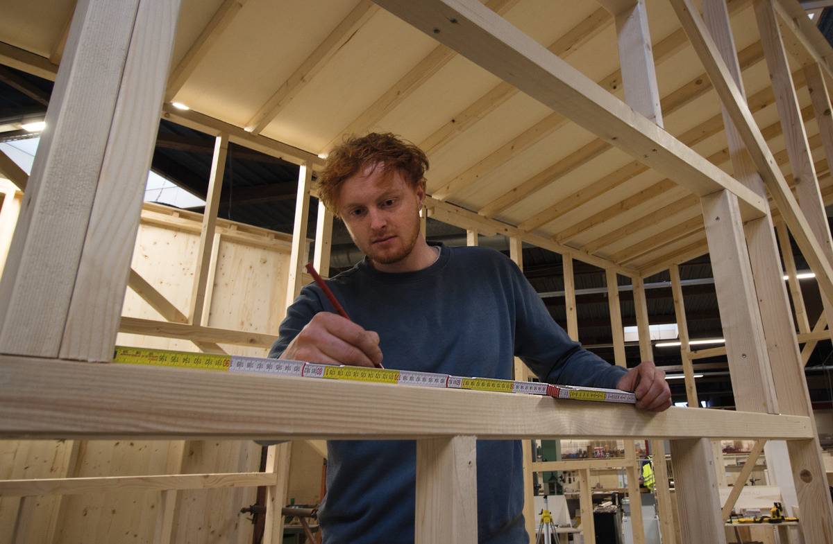 Eine Person misst mit einem Maßband das Holz eines im Bau befindlichen Rahmens in einer Werkstatt.