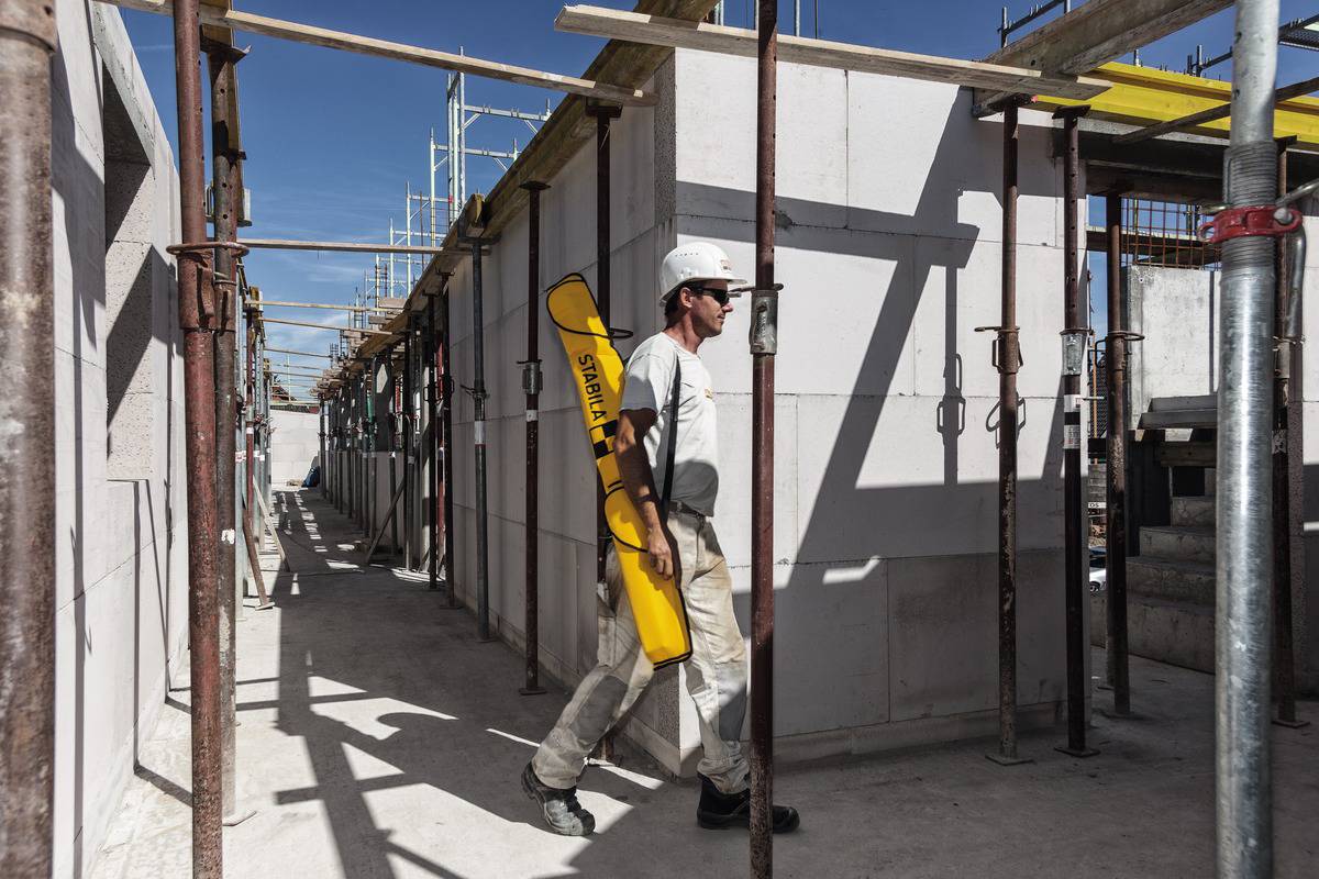 Ein Bauarbeiter geht über eine Baustelle und trägt ein gelbes Werkzeug. Im Hintergrund sind Gerüste und unvollständige Wände zu sehen.