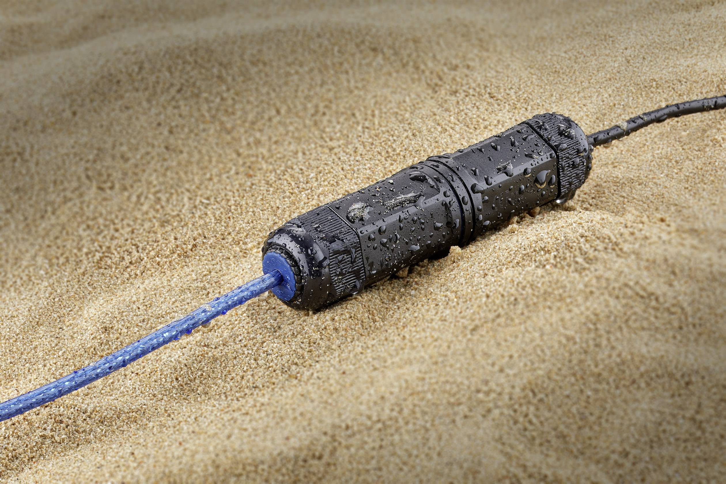Ein wasserdichter Stecker mit blauer Kabelverbindung liegt im Sand. Wassertropfen sind auf der Steckverbindung sichtbar.