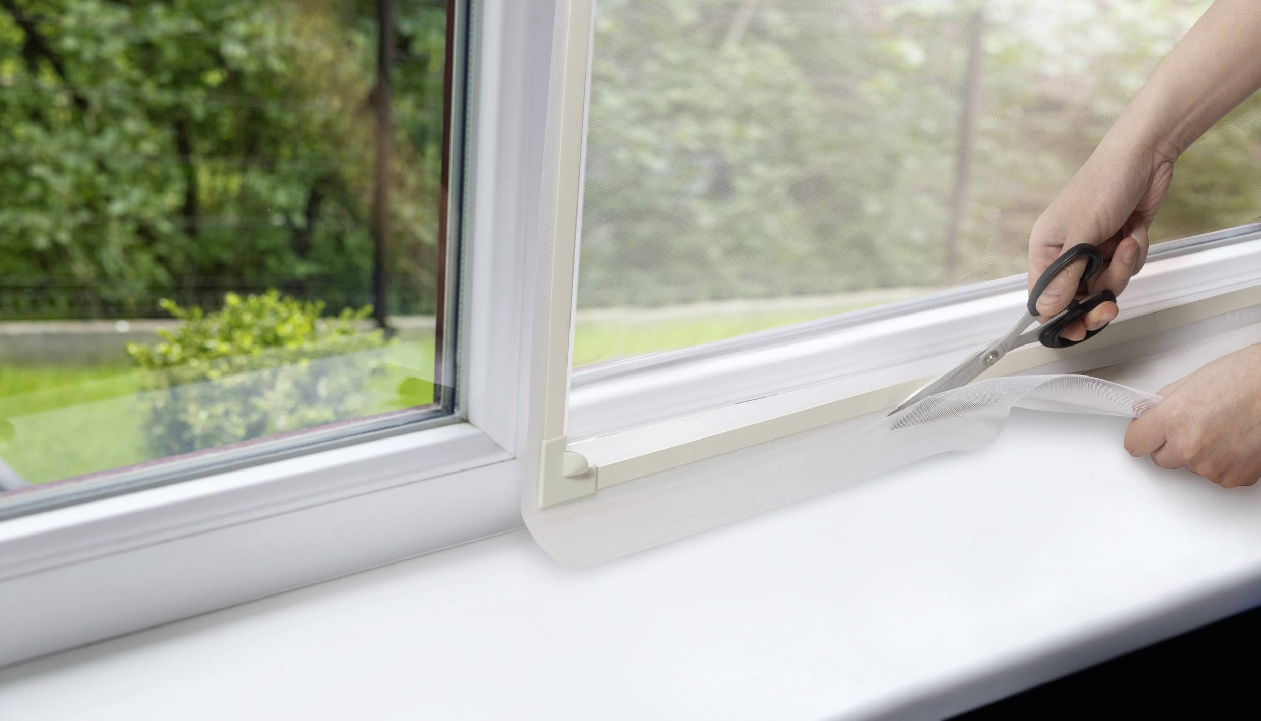 Eine Person schneidet eine durchsichtige Schutzfolie entlang eines Fensterrahmens. Im Hintergrund ist ein Garten sichtbar.