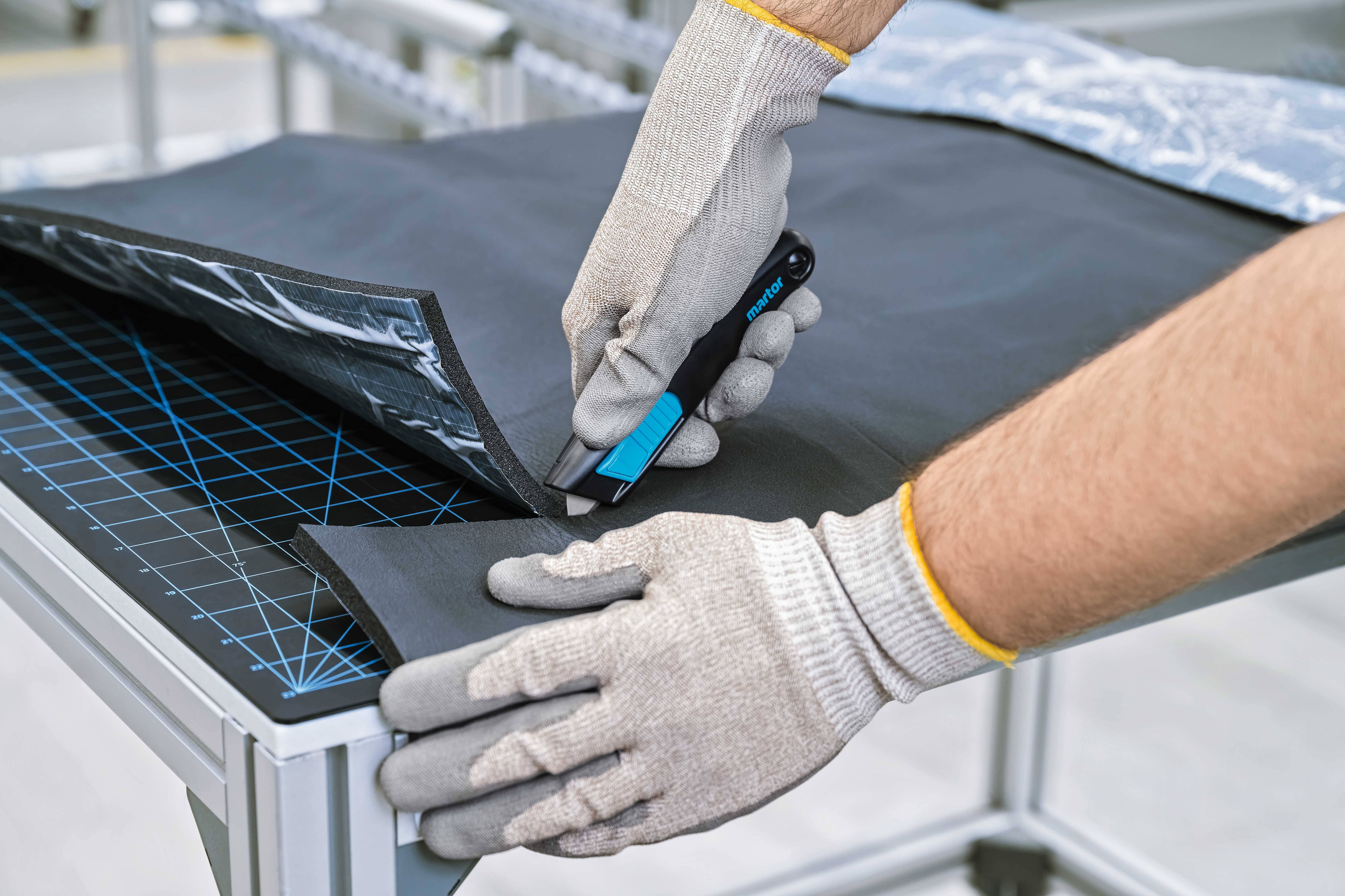 Eine Person schneidet mit einem blauen Cuttermesser ein schwarzes Material auf einem Tisch, trägt dabei Schutzhandschuhe.