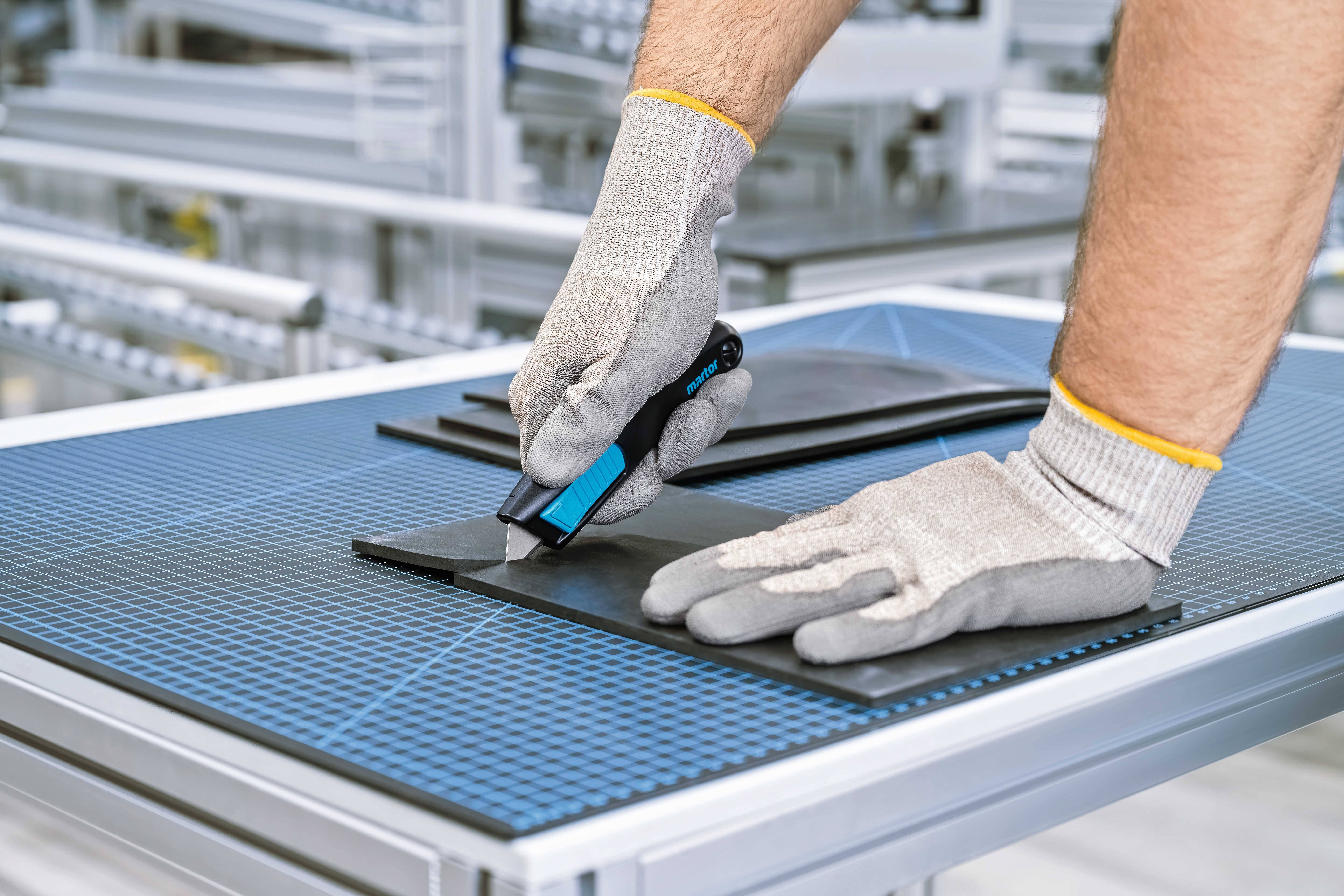 Eine Person mit Handschuhen schneidet schwarze Materialien mit einem Cuttermesser auf einem blauen Schneidetisch in einer Werkstatt.