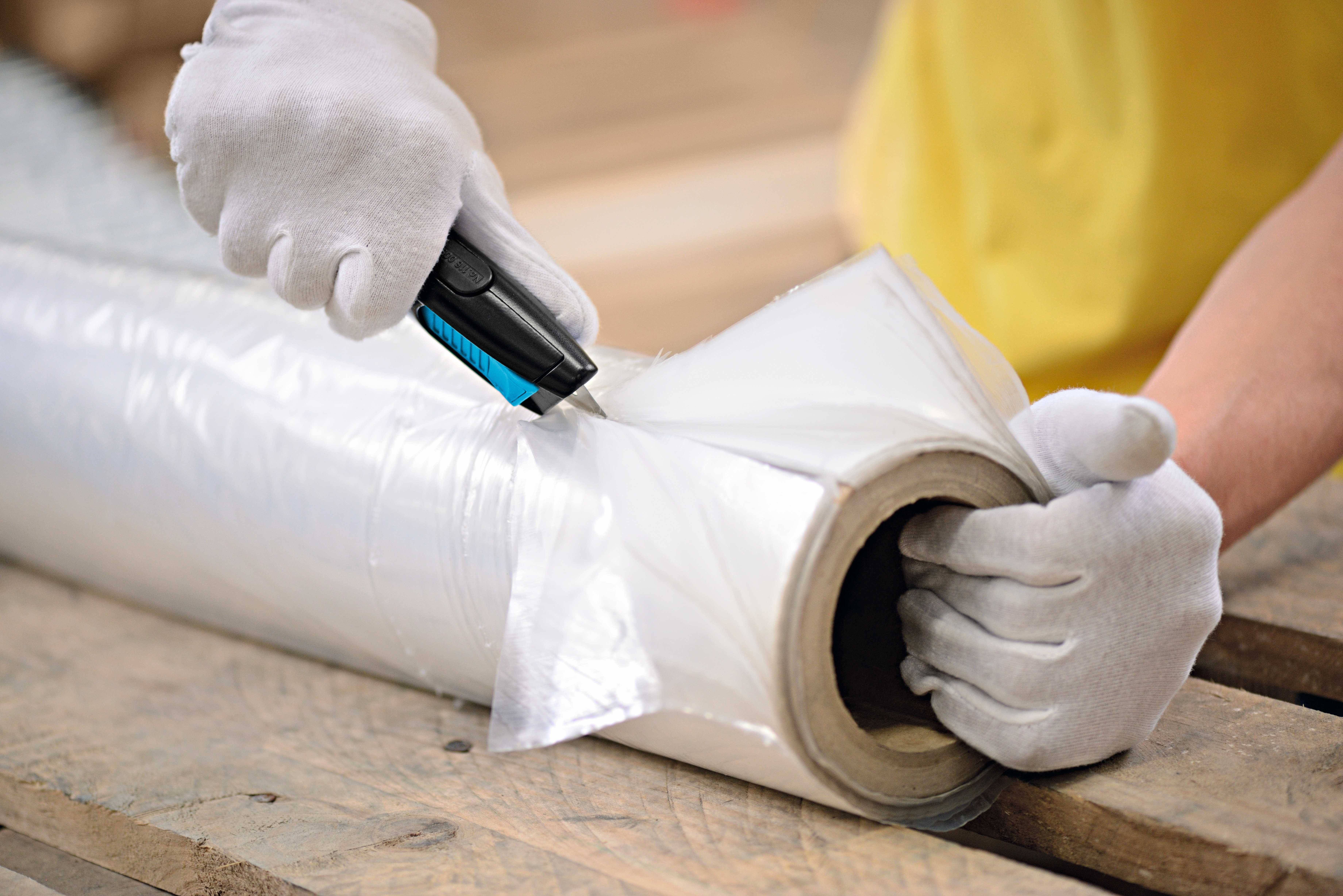 Eine Person trägt weiße Handschuhe und schneidet mit einem Messer Kunststofffolie von einer Rolle auf einer Holzpalette ab.