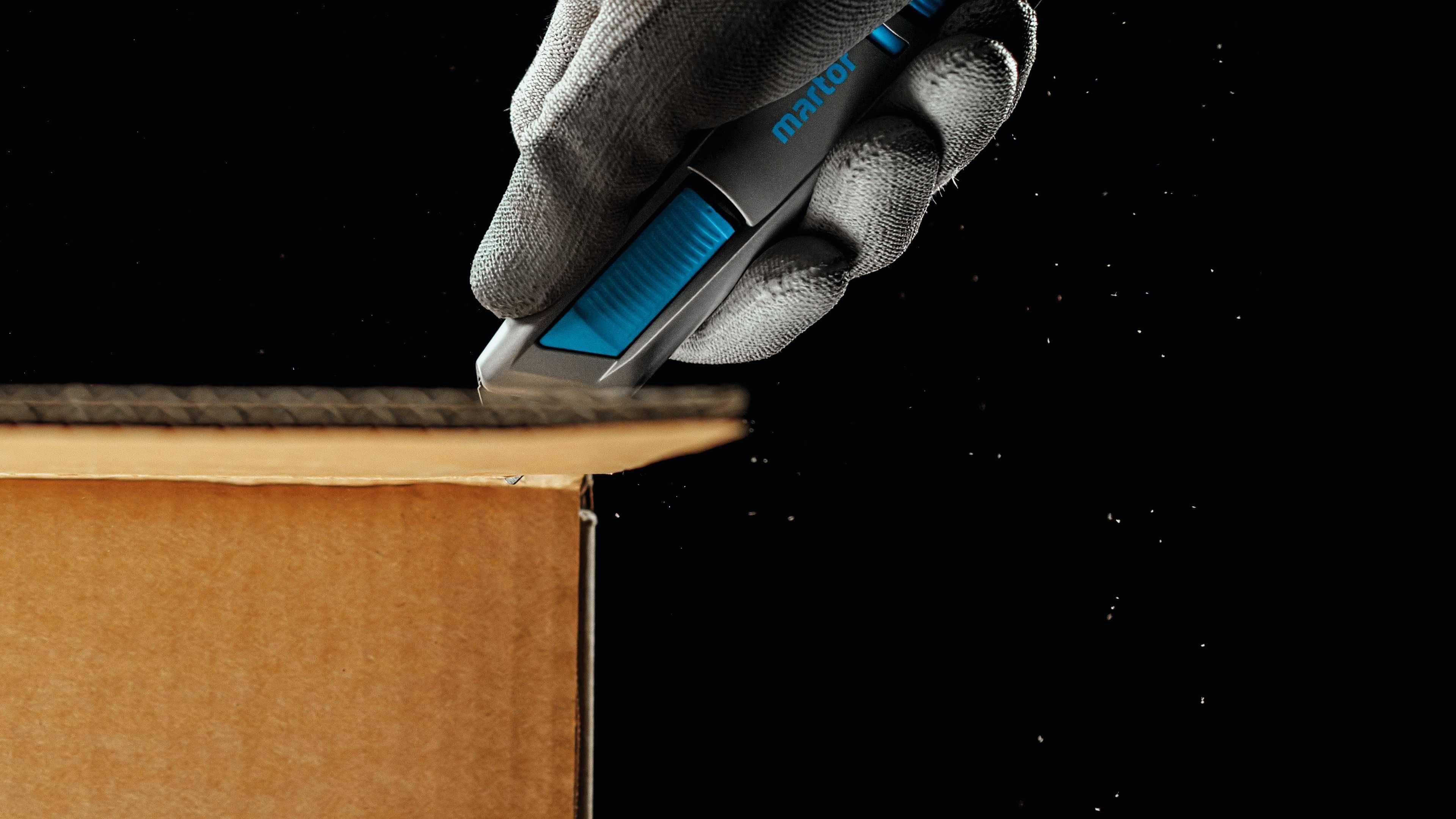 Eine behandschuhte Hand schneidet mit einem blauen Cutter-Messer vorsichtig die Oberseite eines Pappkartons auf schwarzem Hintergrund.