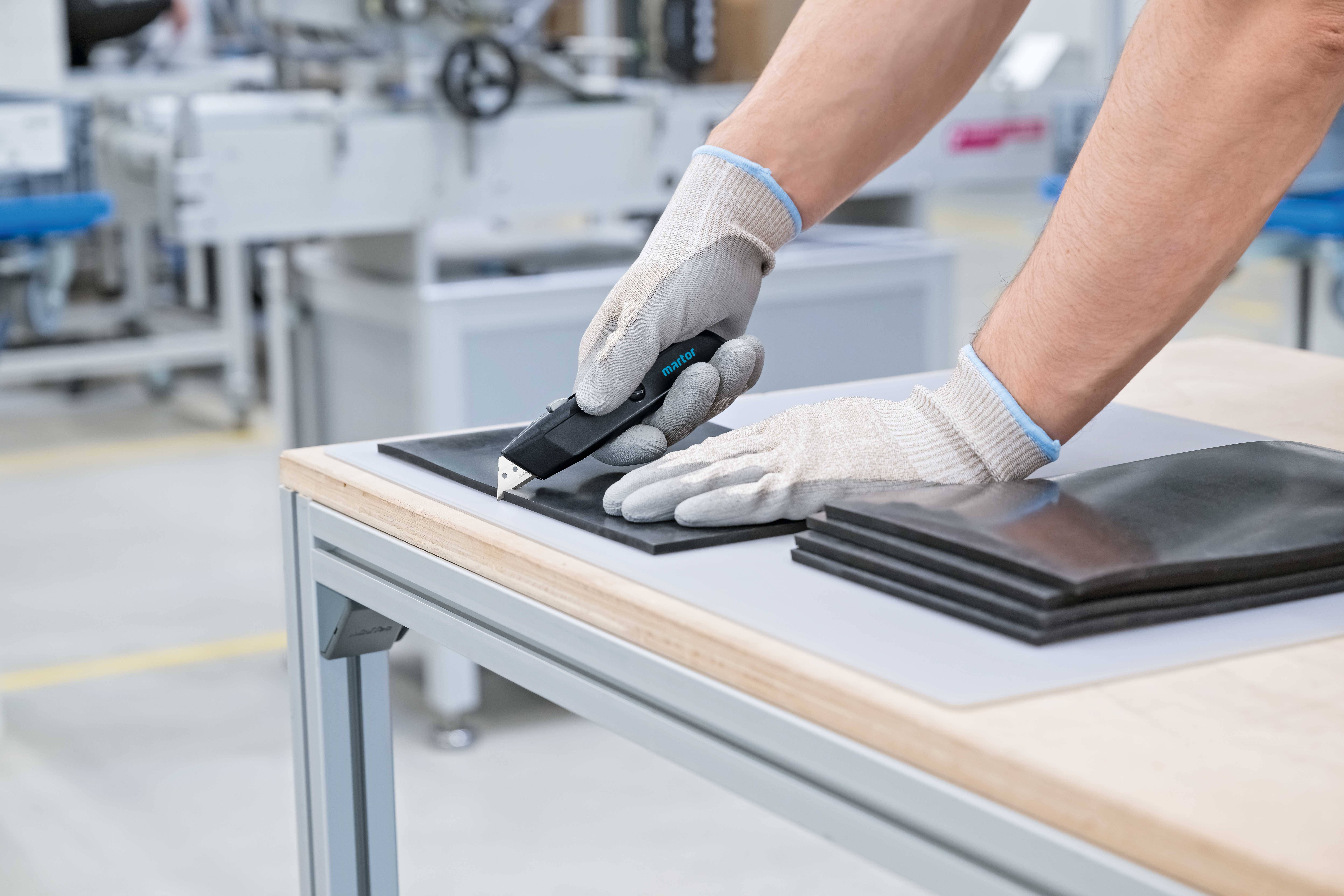 Eine Person mit Handschuhen schneidet mit einem präzisen Werkzeug dünne Materialien auf einem Tisch in einer Werkstatt.