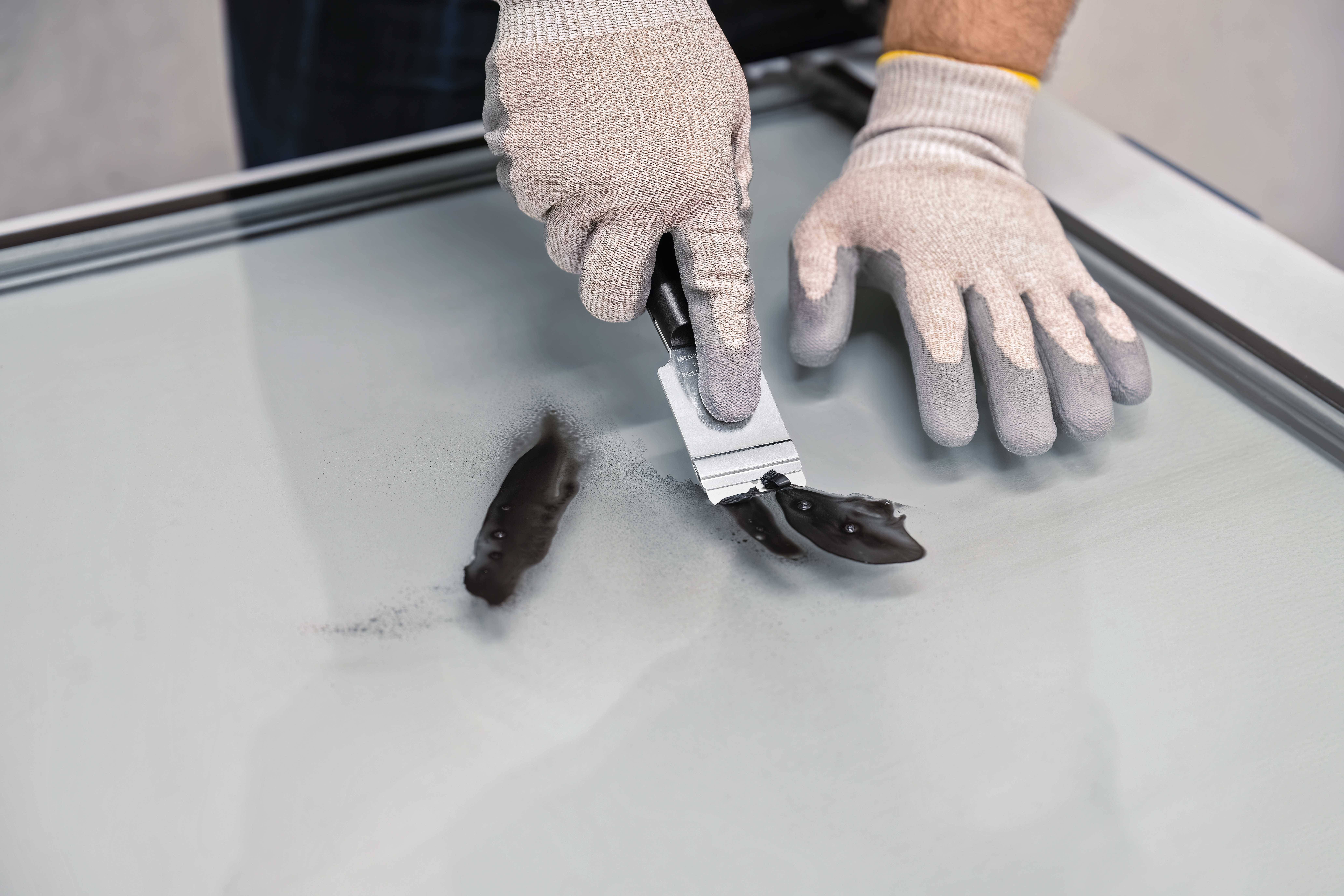 Person mit Handschuhen entfernt schwarzen Schmutz von einer Glasscheibe mit einem Abstreifer.