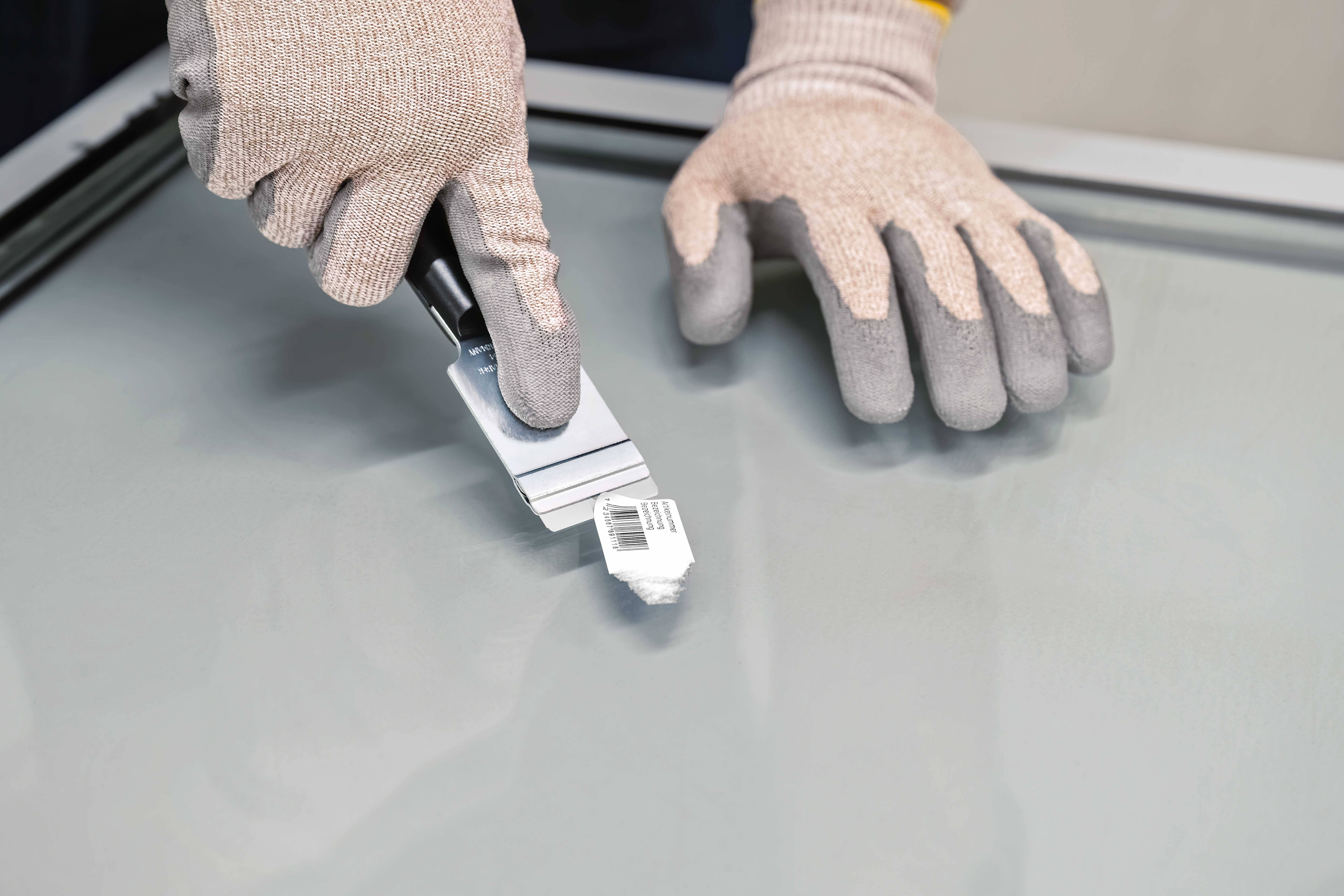 Nahaufnahme von Händen in Arbeitshandschuhen, die mit einem Rasierklingenabstreifer eine glatte Glasoberfläche reinigen.
