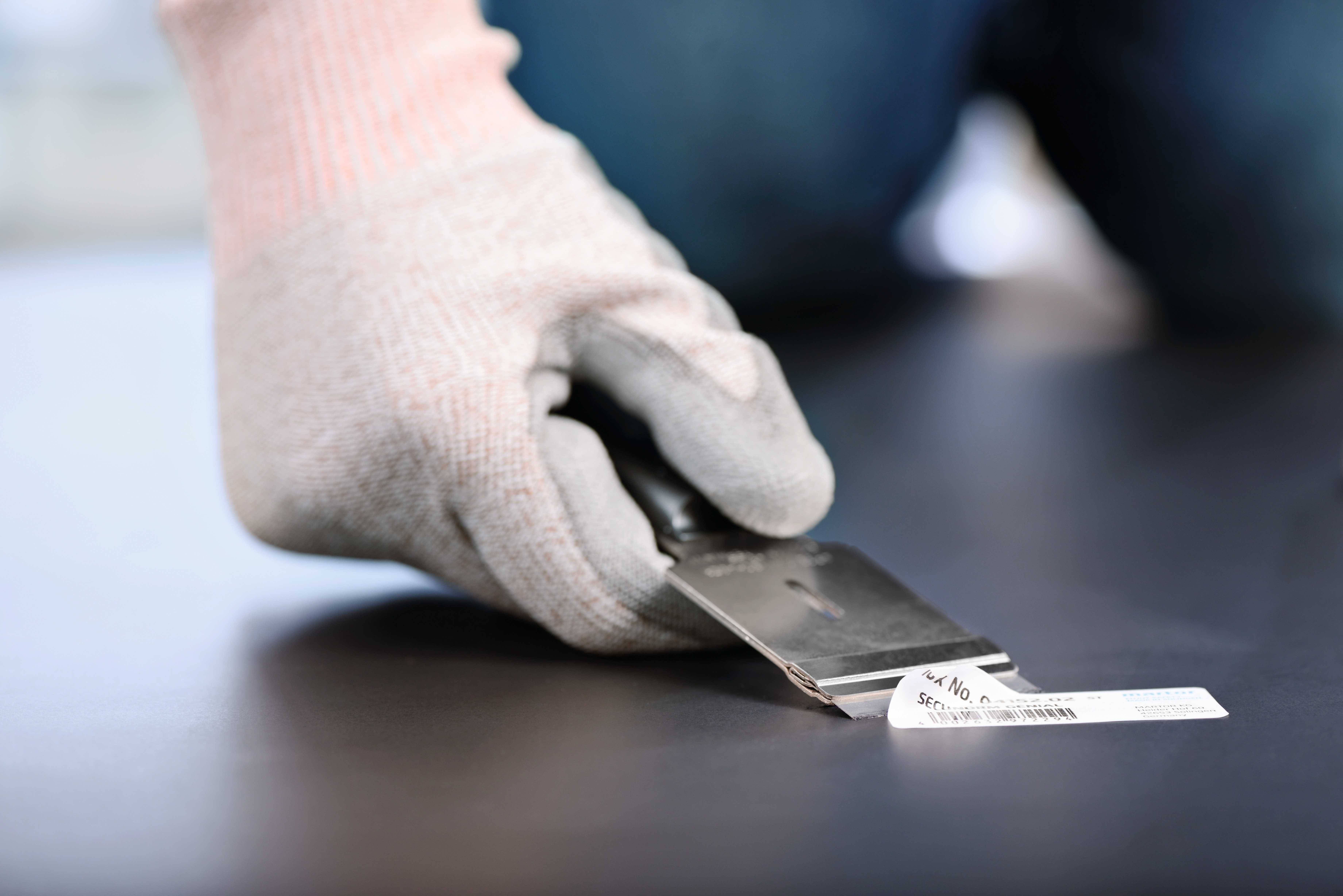 Person mit Schutzhandschuh entfernt Etikett mit Spachtel von einer glatten, dunklen Oberfläche.