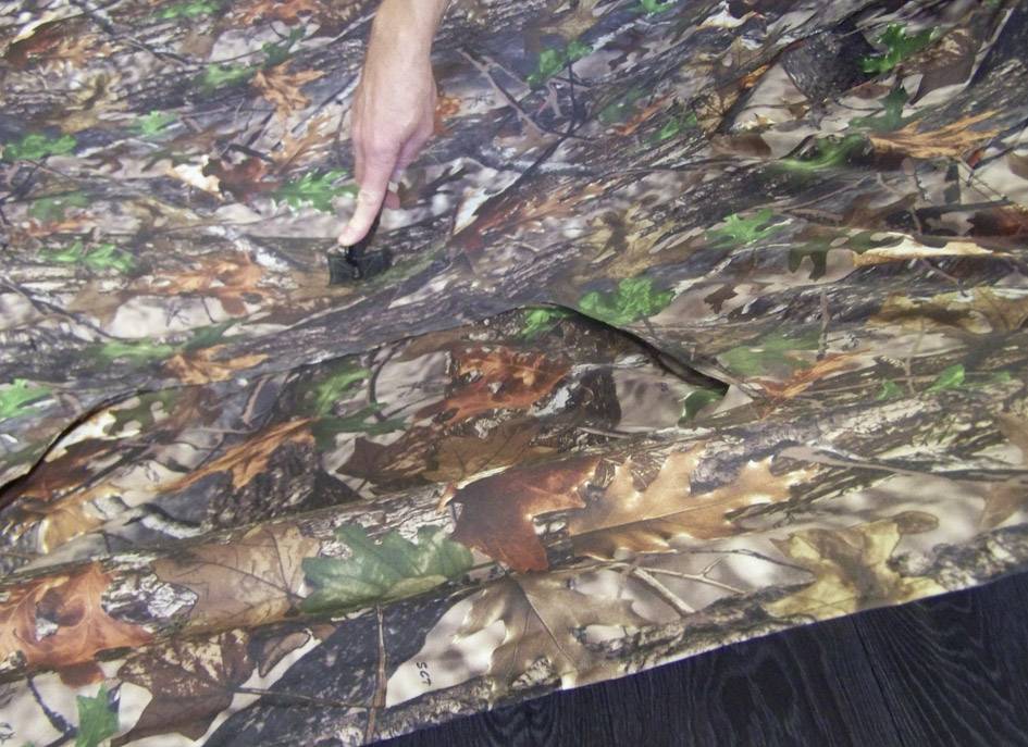 Eine Hand zeigt auf ein Detail eines Tarnnetzes mit Blätter- und Baumrindenmuster, das auf einem dunklen Holzboden liegt.