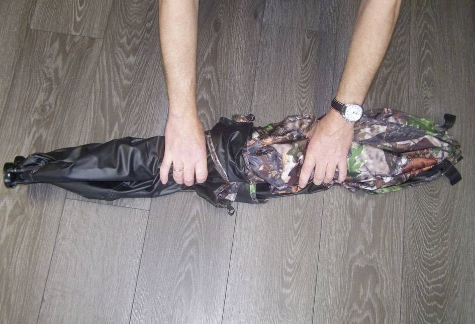 Eine Person rollt ein Tarnzelt auf grauem Holzboden zusammen. Hände sind am Zeltmaterial zu sehen.