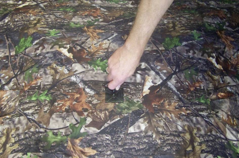 Eine Hand zieht einen Reißverschluss auf einem Tarnmuster-Stoff auf. Der Stoff zeigt ein Waldmuster mit Blättern und Ästen.