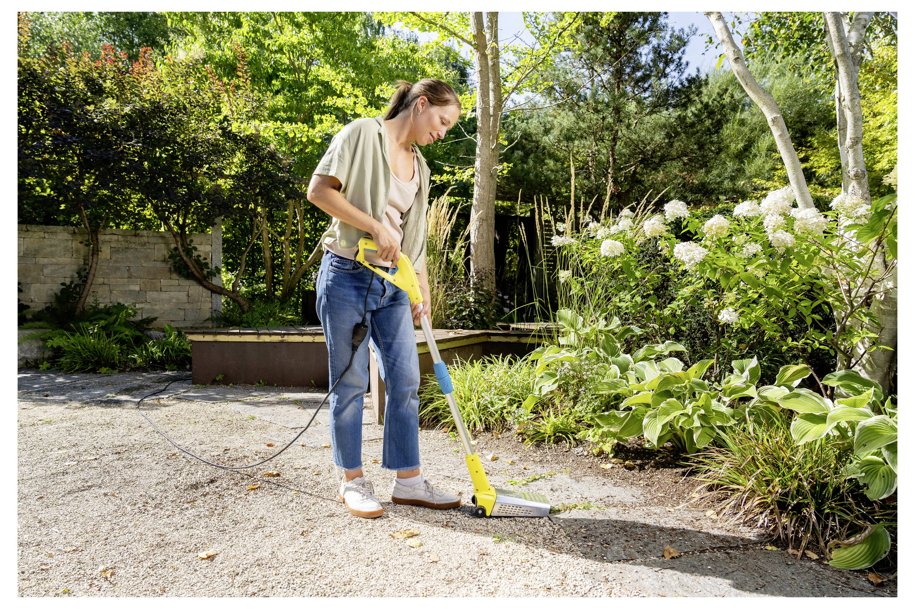 Frau, die einen Hochdruckreiniger verwendet, um eine Terrasse neben einem Garten und blühenden Sträuchern zu reinigen.