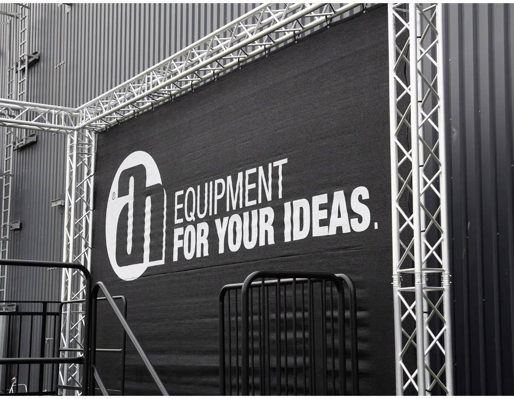 Schwarzes Banner mit weißem Logo 'Equipment for Your Ideas' auf einer Metallgerüststruktur, an einem Gebäude montiert.
